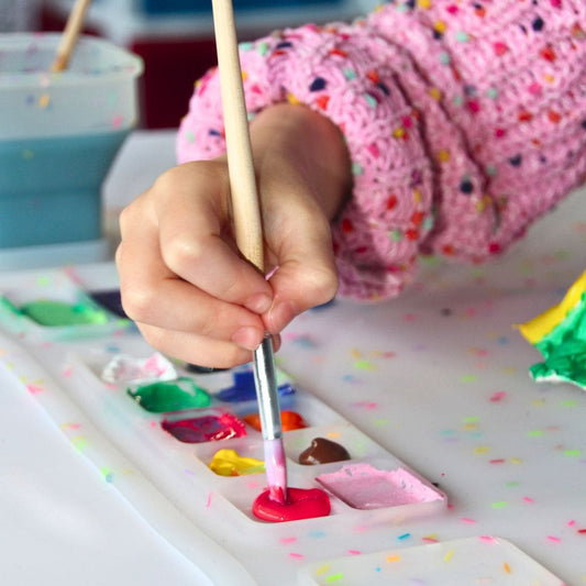 A child's hand using a paintbrush on a silicone painting mat with multi-coloured sprinkles.