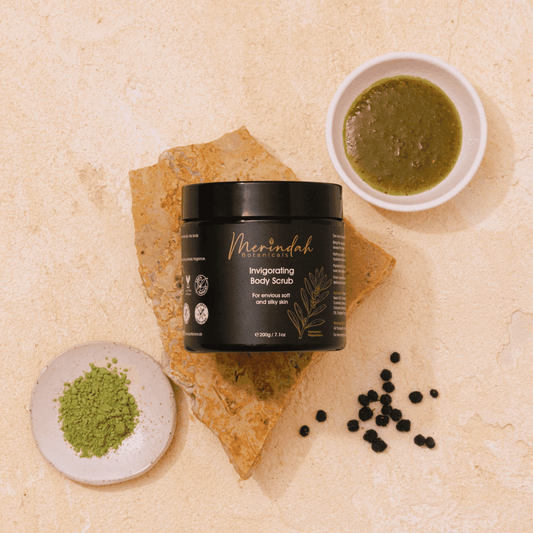 A black jar of Invigorating Body Scrub by Merindah Botanicals is displayed on a stone slab. It is surrounded by small bowls containing exfoliating green powder, green paste, and black peppercorns on a beige textured surface.