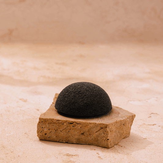 A black, dome-shaped Purifying Konjac Sponge from Merindah Botanicals rests on a textured piece of sandstone against a neutral background. Suitable for all skin types, it elevates your skincare routine with its natural exfoliating touch.