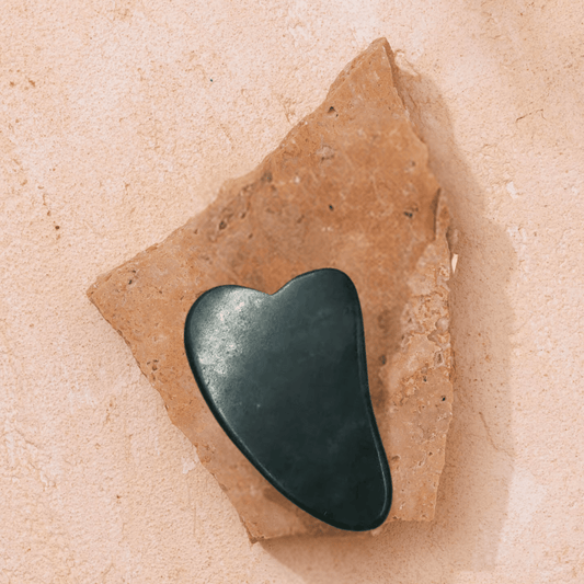 A heart-shaped Gua Sha from Merindah Botanicals sits elegantly on a rugged, irregular piece of beige stone against a complementary textured background, offering a revitalizing facial experience as an integral part of your skincare regimen.