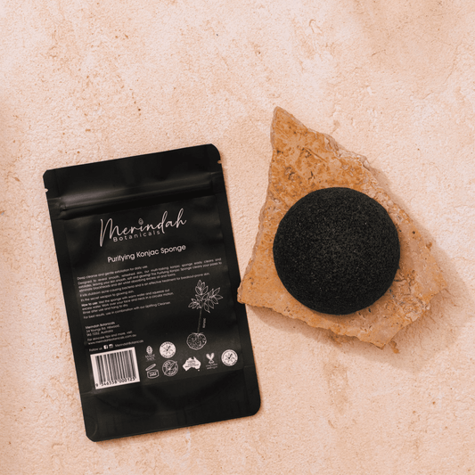 A Purifying Konjac Sponge from Merindah Botanicals, perfect for gentle exfoliation of all skin types, is showcased in its brand packaging on a textured, sand-colored surface. The sponge sits gracefully atop a piece of rough, light brown rock.