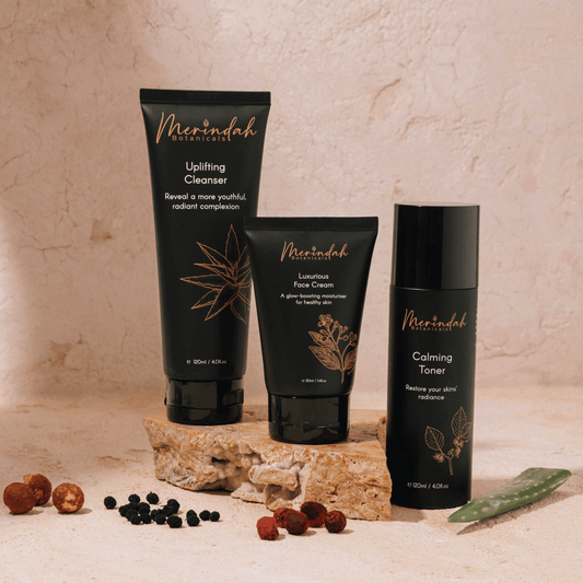 Three products from the Essential Beauty Pack by Merindah Botanicals are displayed on a stone surface. This skincare set includes the Uplifting Cleanser, Luxurious Face Cream, and Calming Toner, each featuring elegant black packaging with botanical illustrations and formulated with natural ingredients.